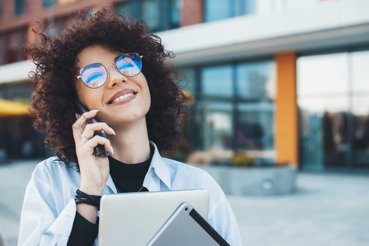 Curly haired lady with glasses is having a phone discussion while posing with her laptopd